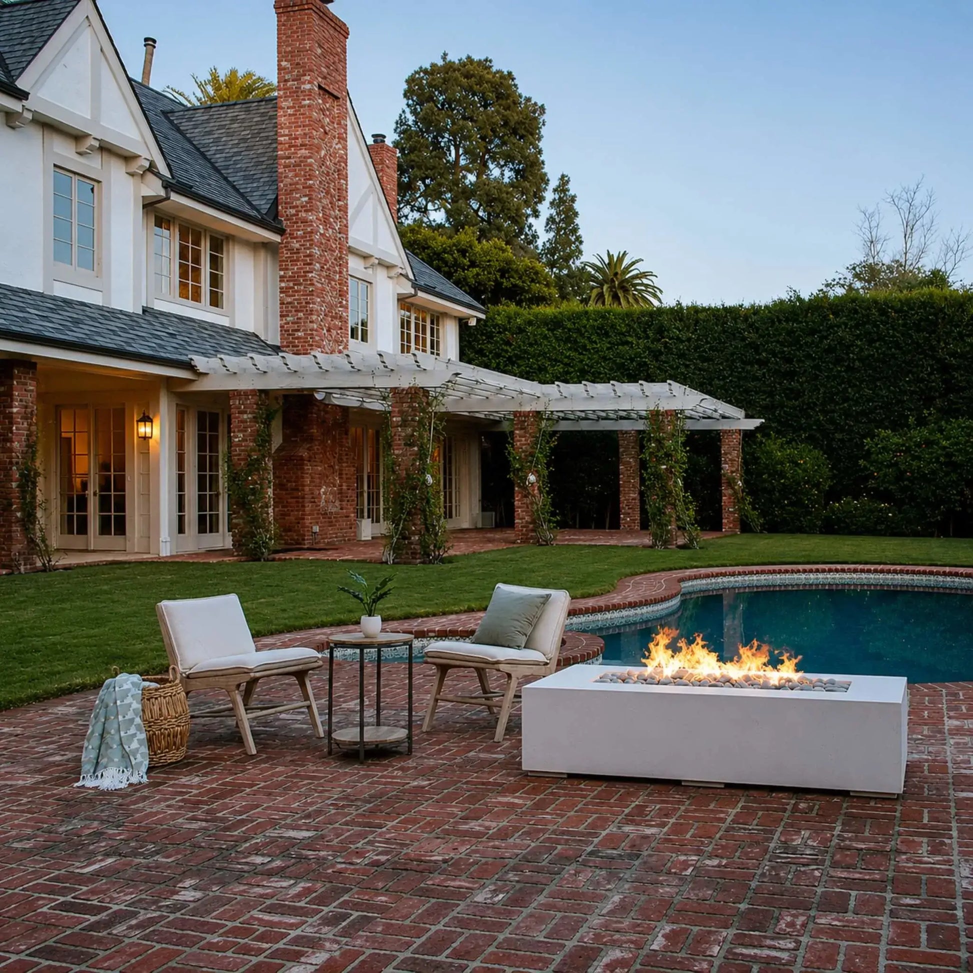 Luxury outdoor entertaining space highlighting the Dekko GFRC Concrete Sonoma 72 x 33 Fire Pit beside a swimming pool and expansive brick patio. Bright linear flames rise from decorative stone media within the rectangular concrete fire pit. Two lounge chairs and a side table overlook the fire feature, while a large traditional home and landscaped grounds provide an elegant backdrop.