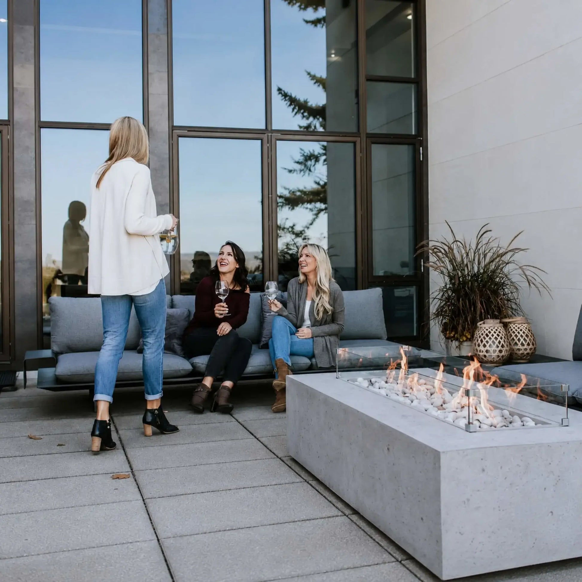 Close-up lifestyle view of the Dekko Sonoma 72 x 33 GFRC Concrete Fire Pit installed on a modern patio outside a contemporary home. Three women enjoy conversation on outdoor sectional seating while the linear fire burns above white decorative stones behind a glass wind guard. The image emphasizes the fire pit’s sleek design and role as a social gathering centerpiece.