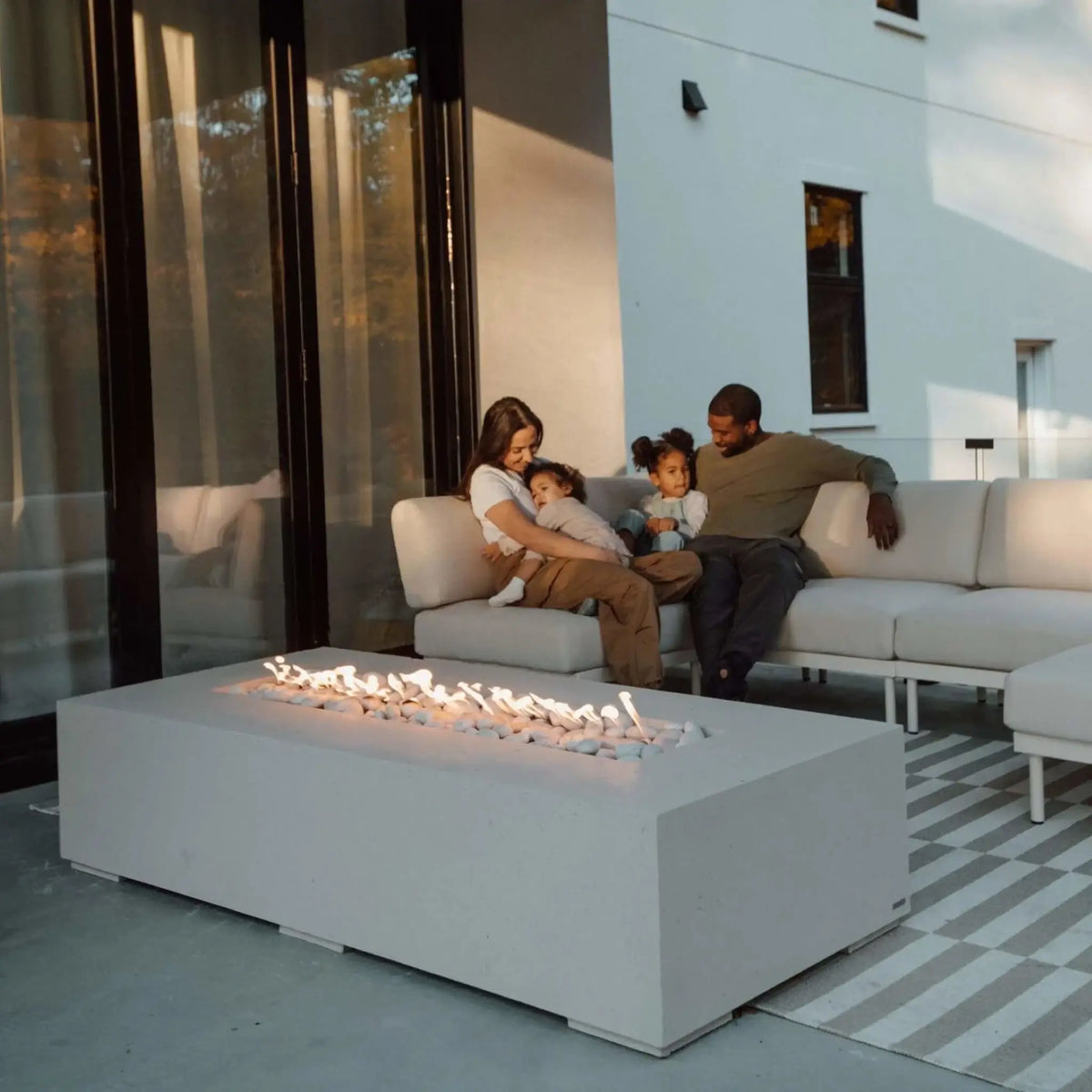 Family gathered around the Dekko GFRC Concrete Sonoma 72 x 33 Fire Pit on a covered modern patio. The large rectangular fire feature displays a continuous linear flame over decorative stones and is positioned in front of comfortable outdoor seating. Parents and children relax together near floor-to-ceiling glass doors, showcasing the fire pit as a family-friendly outdoor living enhancement.