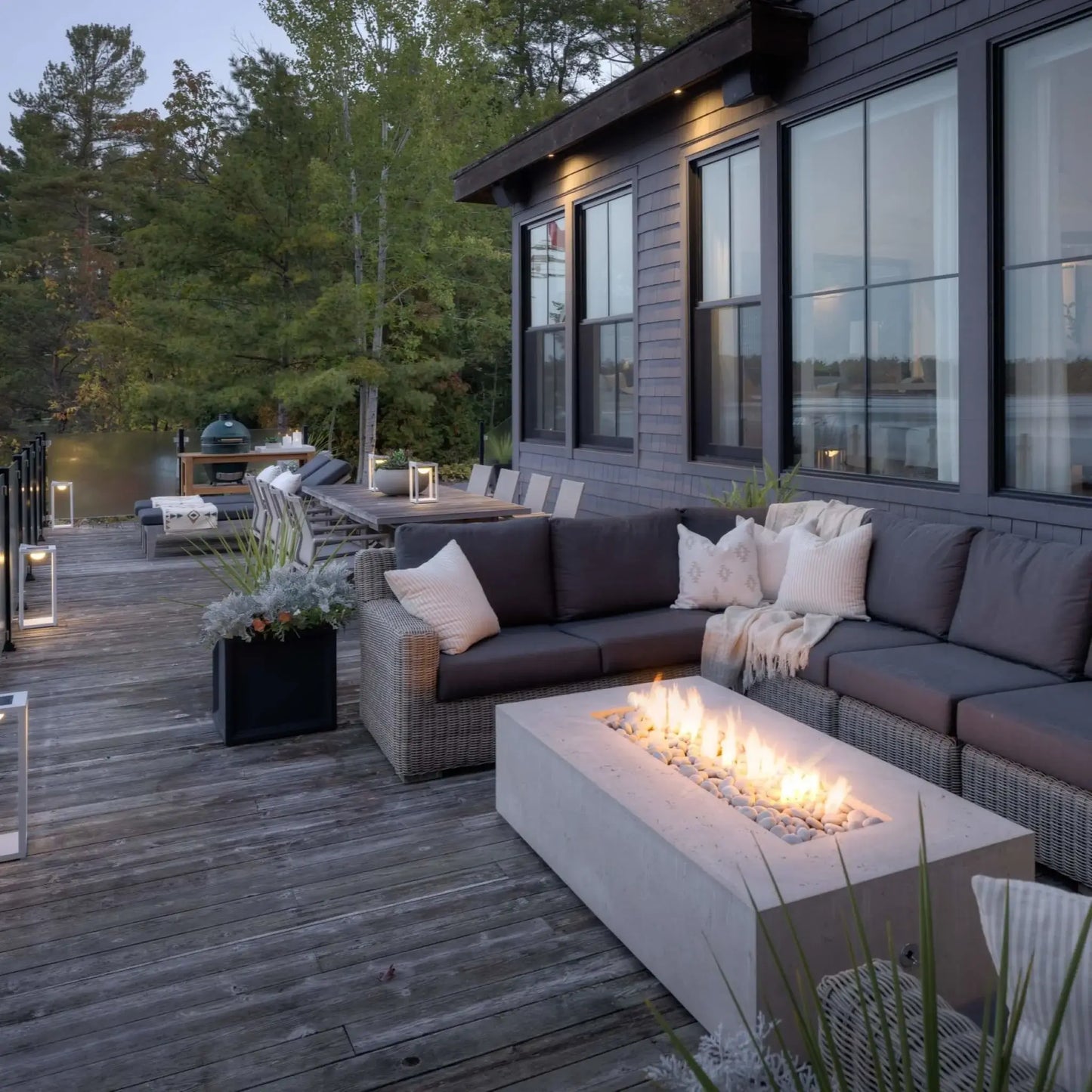 Waterfront deck installation featuring the Dekko Sonoma 72 x 33 GFRC Concrete Fire Pit surrounded by woven outdoor sectional seating. The rectangular concrete fire feature glows with a linear flame display overlooking a calm lake and forested shoreline. Lantern lighting, outdoor dining furniture, and expansive water views create a luxurious lakeside outdoor entertainment setting.