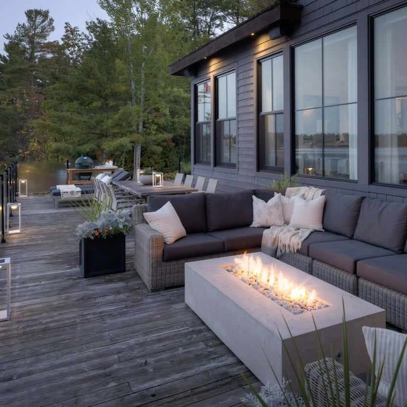 Waterfront deck installation featuring the Dekko Sonoma 72 x 33 GFRC Concrete Fire Pit surrounded by woven outdoor sectional seating. The rectangular concrete fire feature glows with a linear flame display overlooking a calm lake and forested shoreline. Lantern lighting, outdoor dining furniture, and expansive water views create a luxurious lakeside outdoor entertainment setting.