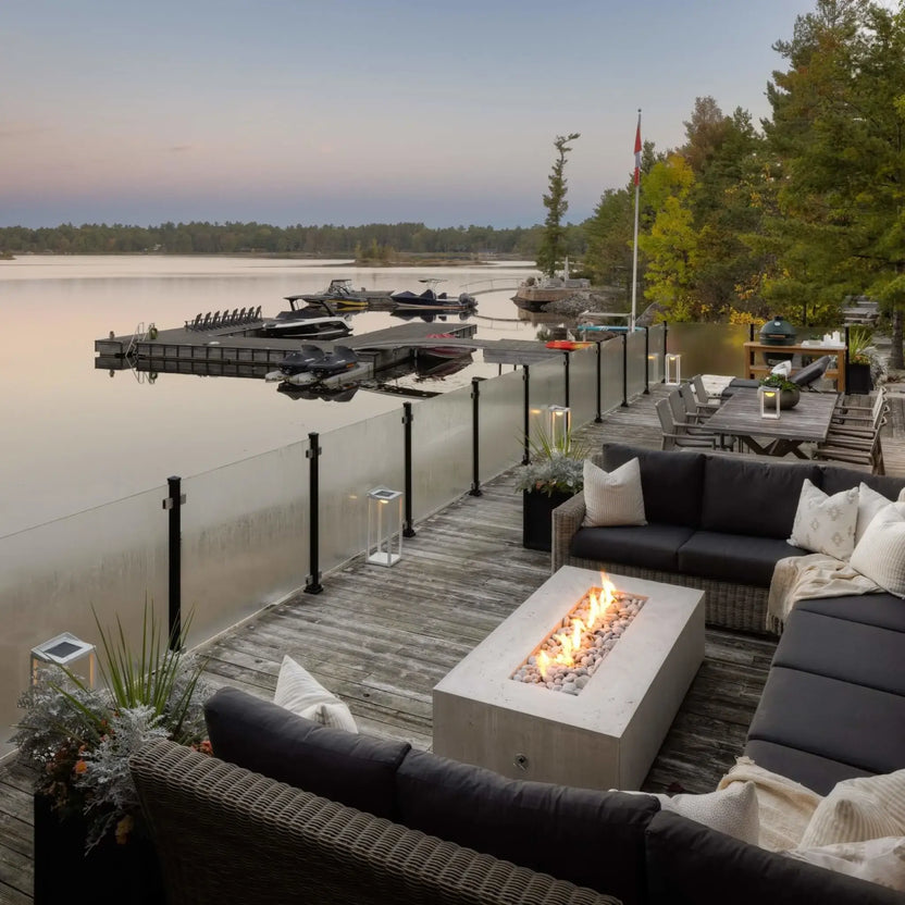 Wide-angle lakeside lifestyle image of the Dekko GFRC Concrete Sonoma 72 x 33 Fire Pit installed on a spacious waterfront deck. The rectangular fire pit burns brightly within a cozy outdoor seating arrangement overlooking docks, boats, and a tranquil lake. Glass railings, modern furnishings, and scenic shoreline views highlight the fire pit’s ability to elevate premium outdoor living spaces.
