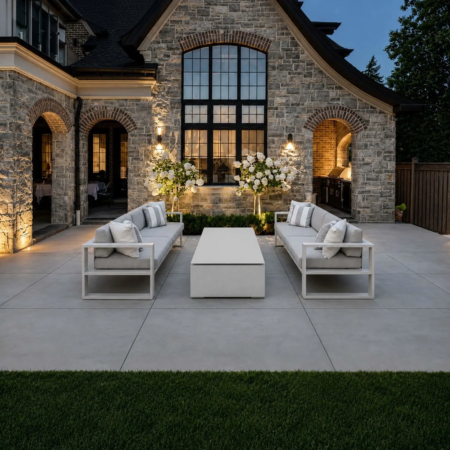 HPC Fire & Hearth Products Controls Dekko GFRC Concrete Sonoma 72 x 33 Fire Pit Table Top Lid showcased in a luxury outdoor courtyard setting. Positioned between two modern cushioned sofas, the rectangular concrete fire pit is covered with its matching tabletop lid, creating a sleek coffee table surface. The elegant patio design, stone home exterior, landscape lighting, and contemporary furnishings highlight the fire pit’s versatility and upscale outdoor appeal.