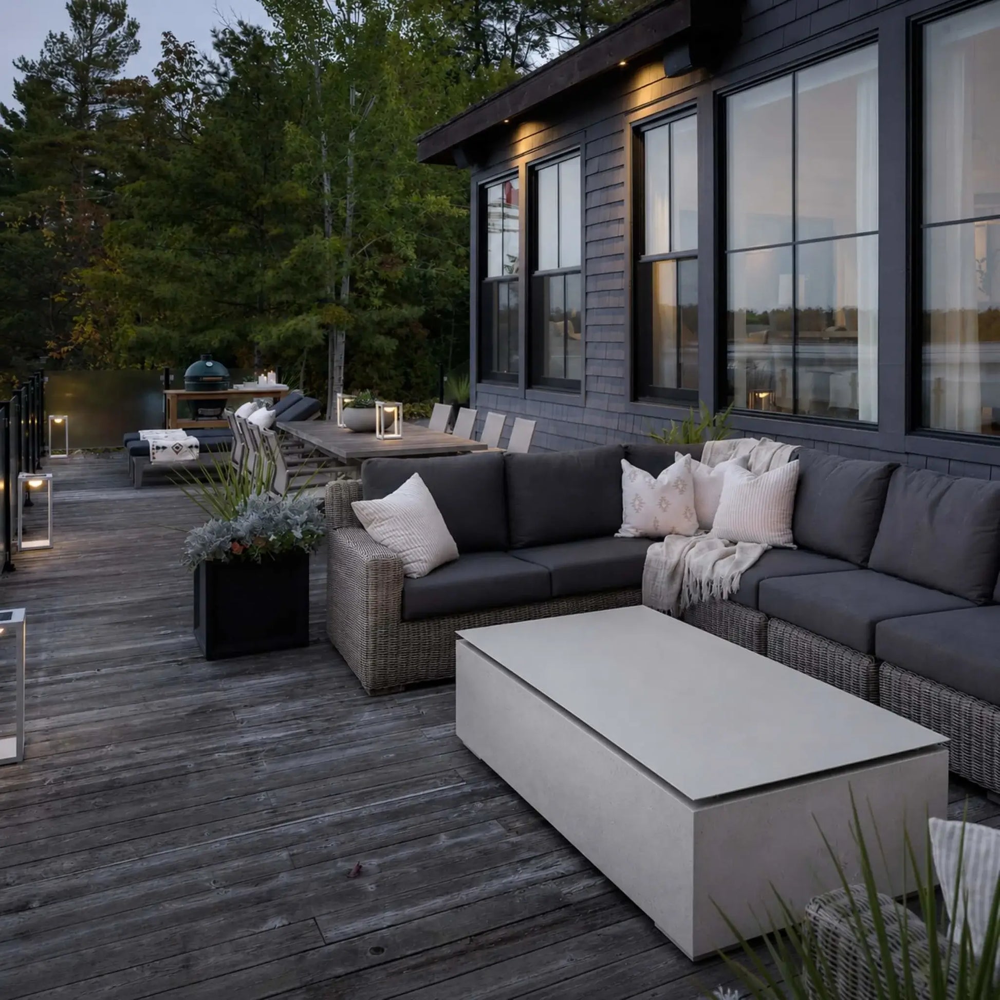 HPC Fire & Hearth Products Controls Dekko GFRC Concrete Sonoma 72 x 33 Fire Pit Table Top Lid displayed on a modern lakeside deck alongside a sectional outdoor sofa. The rectangular GFRC concrete fire pit is covered with its matching tabletop lid, creating a spacious surface for entertaining and relaxation. Surrounded by contemporary outdoor furnishings, warm accent lighting, and natural wooded views, the fire pit serves as a stylish focal point for outdoor living.