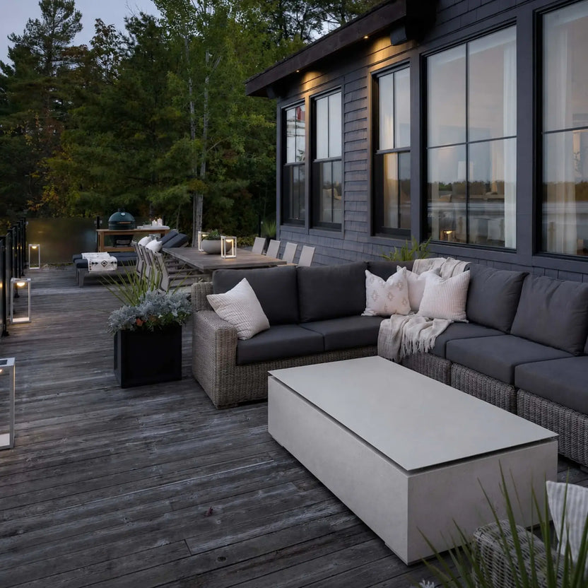 HPC Fire & Hearth Products Controls Dekko GFRC Concrete Sonoma 72 x 33 Fire Pit Table Top Lid displayed on a modern lakeside deck alongside a sectional outdoor sofa. The rectangular GFRC concrete fire pit is covered with its matching tabletop lid, creating a spacious surface for entertaining and relaxation. Surrounded by contemporary outdoor furnishings, warm accent lighting, and natural wooded views, the fire pit serves as a stylish focal point for outdoor living.