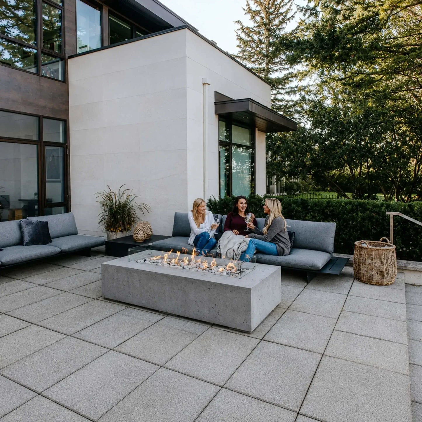 HPC Fire Hearth Products Controls Dekko GFRC Concrete Sonoma 72 x 33 Fire Pit Tempered Glass Wind Guard Lifestyle 2 installation featuring a modern outdoor patio with gray sectional seating, decorative fire stones, and a linear gas flame. Three women relax and socialize around the rectangular concrete fire pit in front of a contemporary luxury home, showcasing the fire table as a functional and stylish outdoor gathering centerpiece.