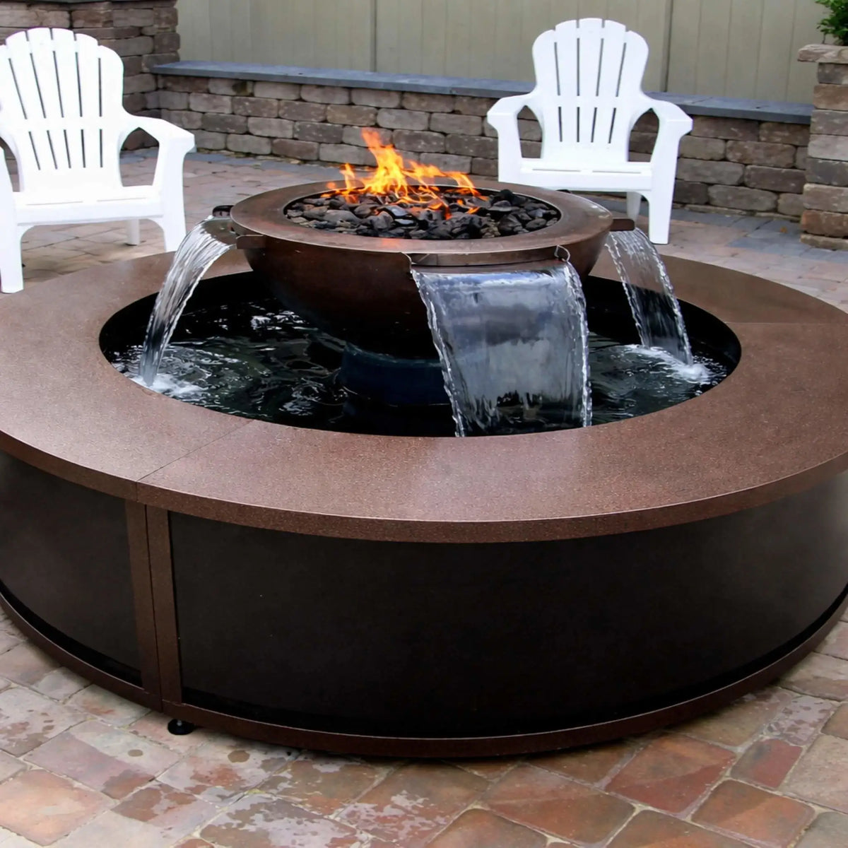Outdoor patio installation of the HPC Fire Evolution 360 fire and water feature with a round table-style enclosure. Water flows evenly from multiple scuppers into the surrounding basin while a gas flame burns above black lava rock. White Adirondack chairs and a stone retaining wall create a relaxed backyard setting.