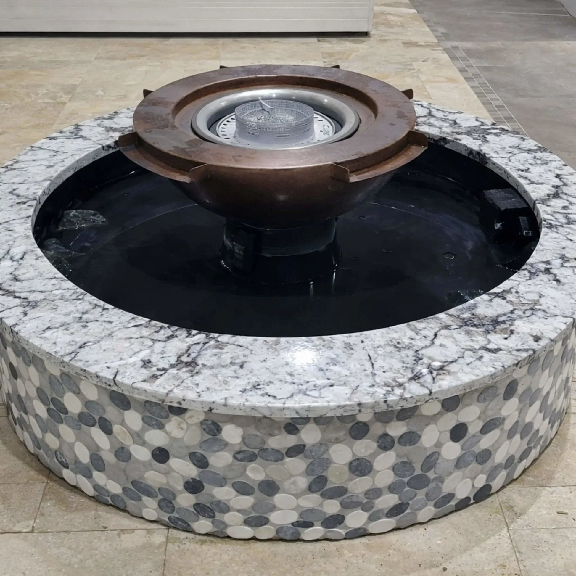 Installed Evolution 360 fire water feature surrounded by a circular stone and tile enclosure. The copper bowl sits elevated in the center with water basin below, showcasing a modern integrated fire pit and fountain design. The unit is shown unlit, emphasizing structure, finish materials, and installation detail.