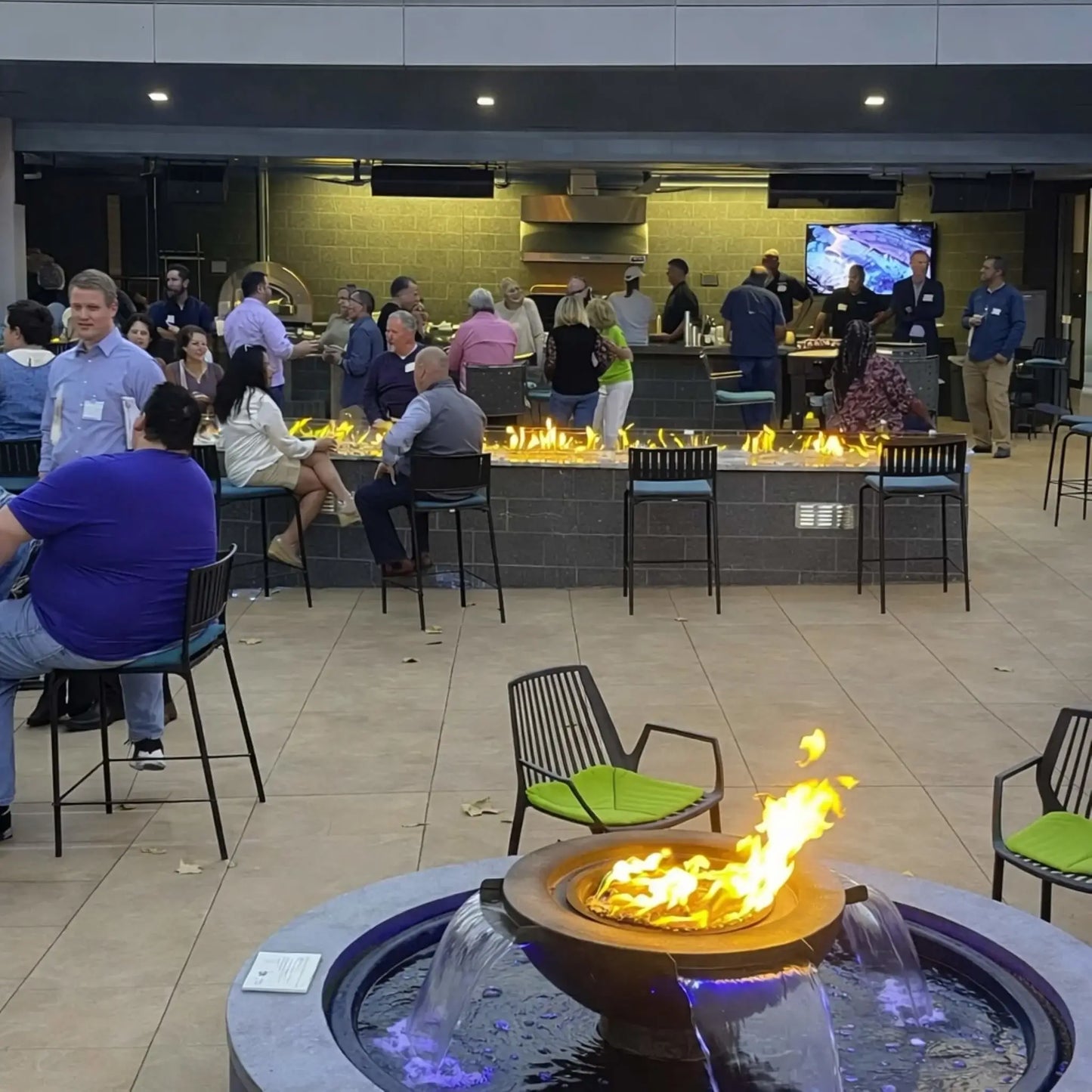 Commercial outdoor lounge space featuring the HPC Fire Evolution 360 fire and water feature in the foreground, with flames and cascading water illuminated by blue LED lighting. Guests gather around a long linear fire feature and bar area in the background, highlighting a hospitality-grade fire and water installation.