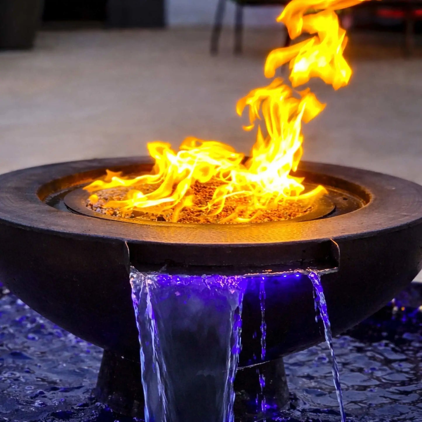Close-up of the Evolution 360 fire and water feature showing bright yellow flames dancing over fire media while water pours smoothly from a scupper below. Blue LED lighting enhances the cascading water effect, emphasizing the combination of fire and water elements in this HPC fire feature system.
