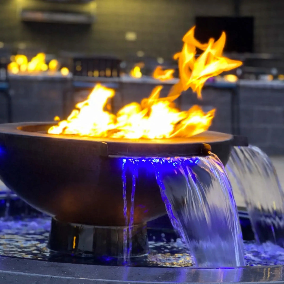 Detailed side view of the Evolution 360 fire and water feature with flames rising above the bowl and water cascading from dual scuppers into the basin. Blue LED lighting highlights the water flow, creating a dramatic contrast between fire and water in this premium HPC fire feature design.