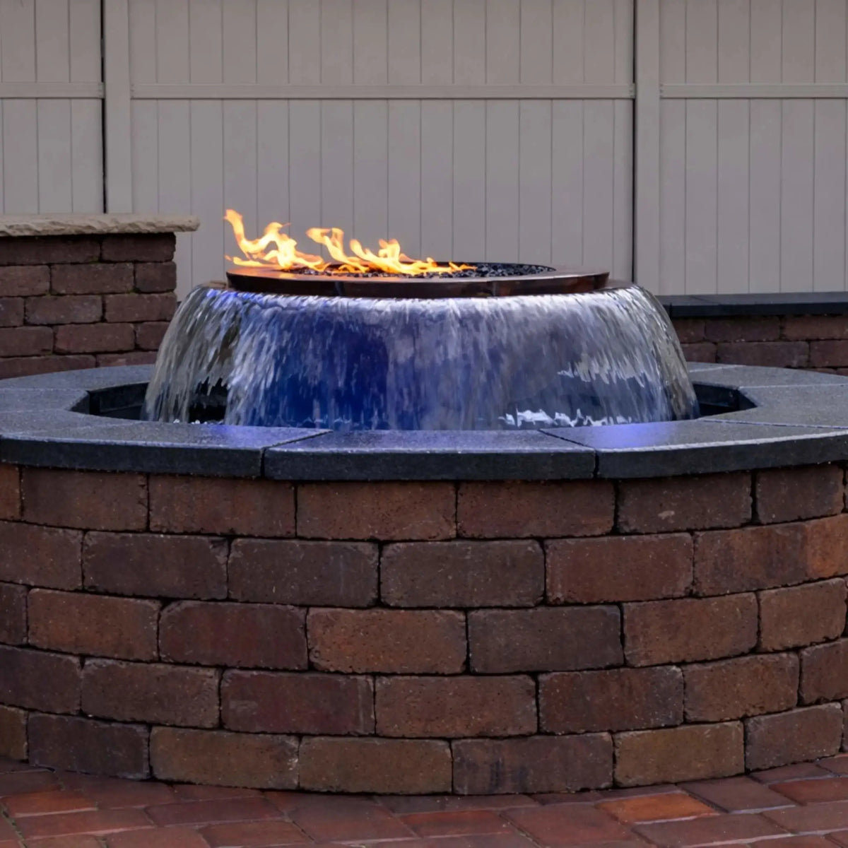 A built-in HPC Fire Evolution 360 True Fire Water feature installed in a circular brick enclosure, showing a smooth 360-degree waterfall flowing evenly around the bowl with a linear flame burning above. The image highlights the clean stone cap, backyard privacy fence, and the seamless integration of fire and water in a residential patio setting.