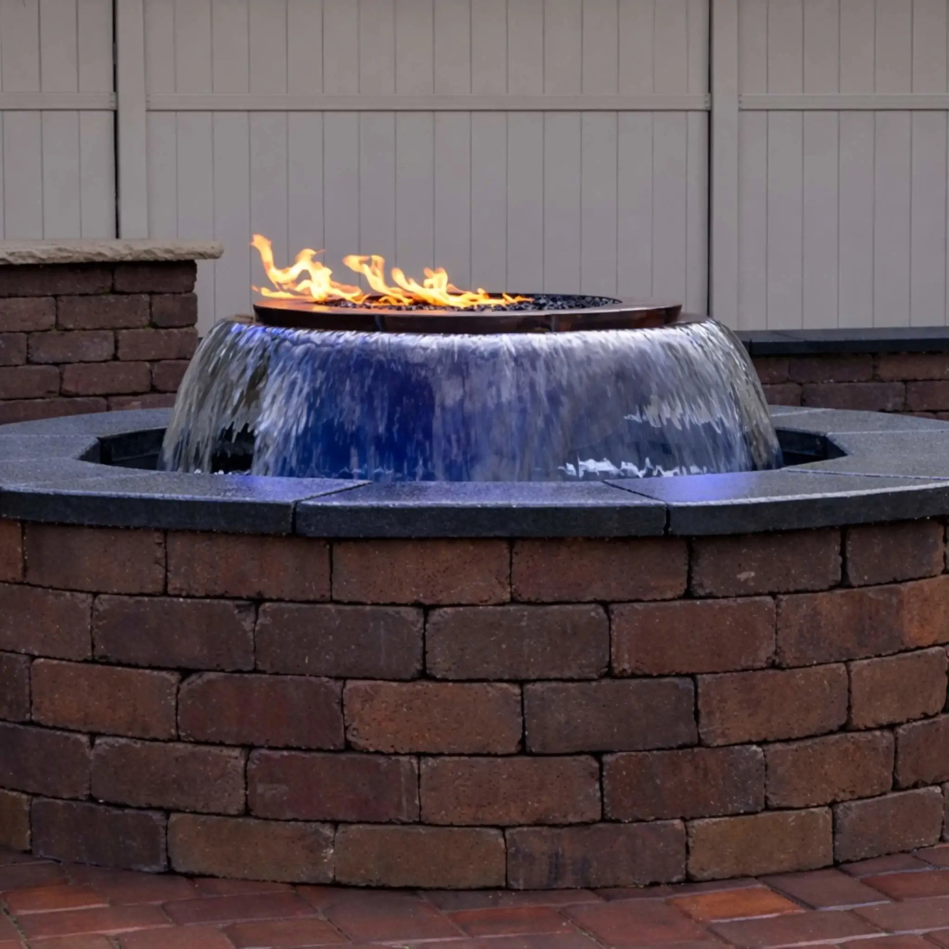 A built-in HPC Fire Evolution 360 True Fire Water feature installed in a circular brick enclosure, showing a smooth 360-degree waterfall flowing evenly around the bowl with a linear flame burning above. The image highlights the clean stone cap, backyard privacy fence, and the seamless integration of fire and water in a residential patio setting.
