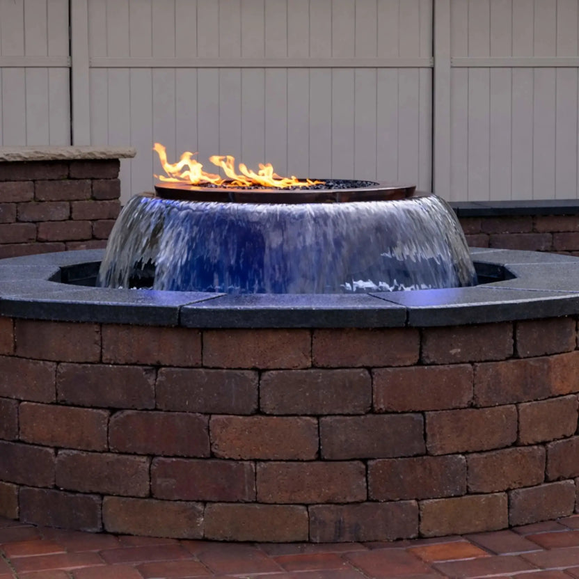A built-in HPC Fire Evolution 360 True Fire Water feature installed in a circular brick enclosure, showing a smooth 360-degree waterfall flowing evenly around the bowl with a linear flame burning above. The image highlights the clean stone cap, backyard privacy fence, and the seamless integration of fire and water in a residential patio setting.