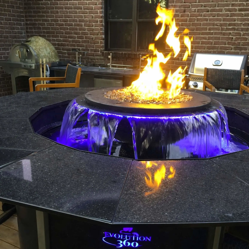 Premium outdoor kitchen setting featuring the Evolution 360 True Fire Water feature integrated into a polished stone countertop. Flames rise above fire glass while water cascades below with blue LED lighting, surrounded by a grill, pizza oven, and brick wall backdrop.