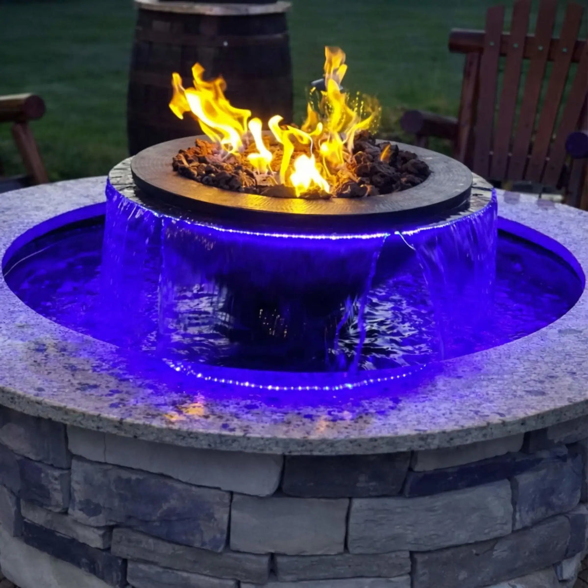 Evening backyard scene featuring the HPC Fire Evolution 360 True Fire Water feature with bright flames over lava rock and a smooth circular waterfall glowing with blue LED lighting. The stone enclosure and surrounding wooden chairs create a warm, inviting patio atmosphere.