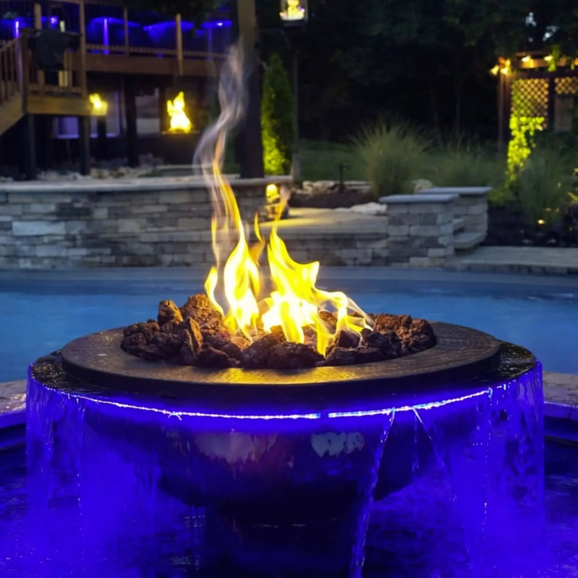 Close-up nighttime view of the Evolution 360 True Fire Water feature with tall flames rising above dark fire media. Water cascades evenly around the bowl, illuminated by blue LED lighting, with a softly lit outdoor living space and deck in the background.