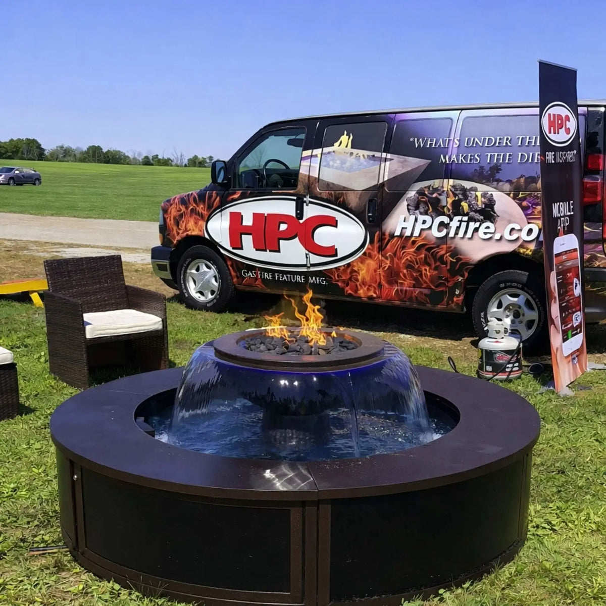 Outdoor demo setup of the HPC Fire Evolution 360 fire and water feature placed on grass, with flames burning above the central bowl and water flowing around it. An HPC-branded van and banner in the background highlight the product in a live event or showcase setting.