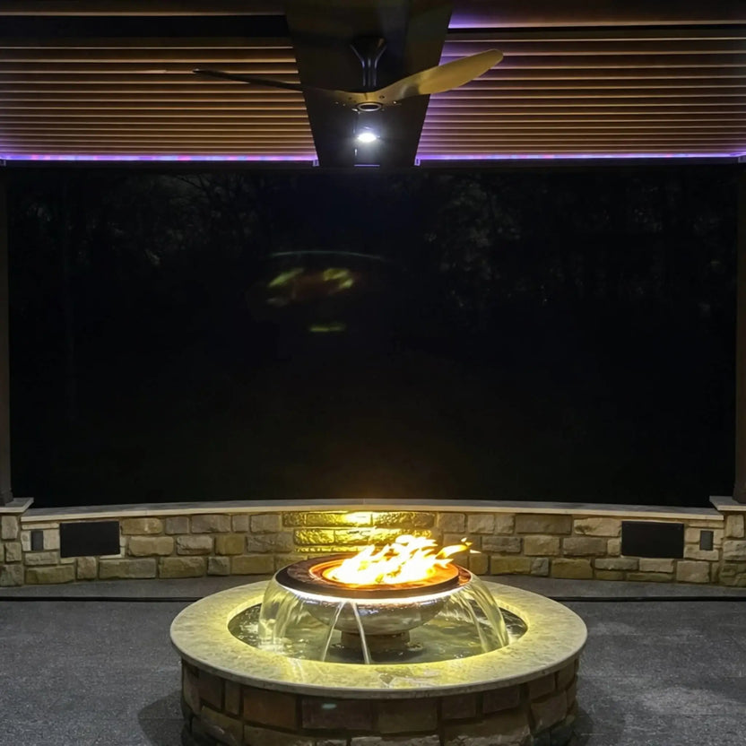 Covered patio installation of the Evolution 360 True Fire Water feature glowing with warm lighting beneath a pergola ceiling. Flames burn steadily above the bowl while water cascades below, creating a cozy nighttime focal point framed by stone walls and outdoor lighting.