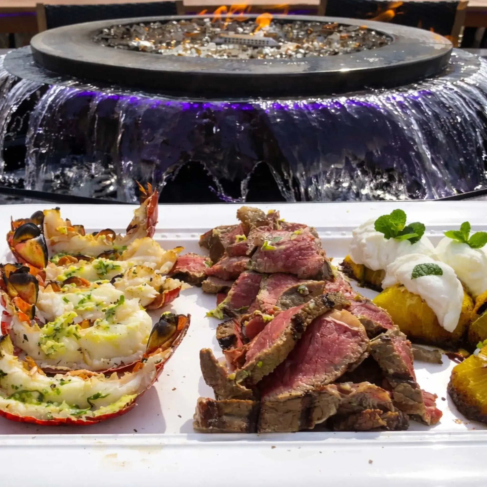 Close-up dining scene with a plated meal of lobster, sliced steak, and roasted vegetables in the foreground, set in front of the Evolution 360 fire and water feature. The cascading water and blue LED lighting add ambiance to an upscale outdoor dining experience.