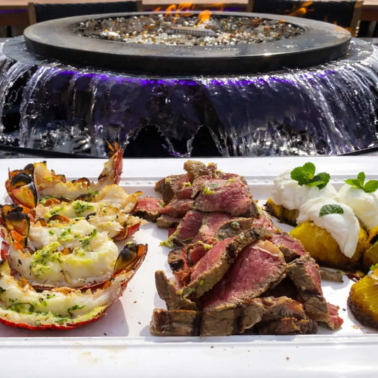 Close-up dining scene with a plated meal of lobster, sliced steak, and roasted vegetables in the foreground, set in front of the Evolution 360 fire and water feature. The cascading water and blue LED lighting add ambiance to an upscale outdoor dining experience.