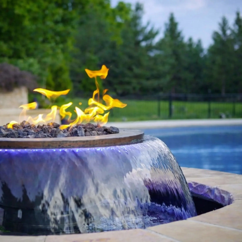 Side angle view of the Evolution 360 True Fire Water feature near a pool, showing flames dancing above fire media while water flows smoothly over the bowl edge. Blue LED lighting enhances the waterfall effect against a landscaped backyard setting.