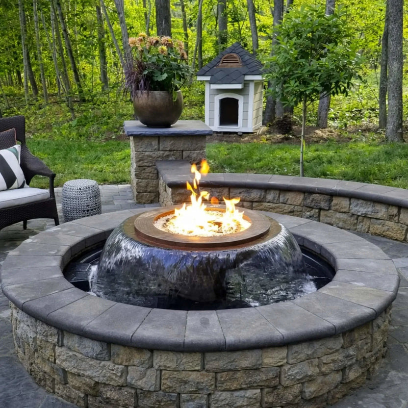 Backyard patio with a built-in Evolution 360 fire and water feature surrounded by curved stone seating walls. Flames rise from the center while water cascades into the basin, with trees, plants, and a small outdoor structure completing the serene landscape.