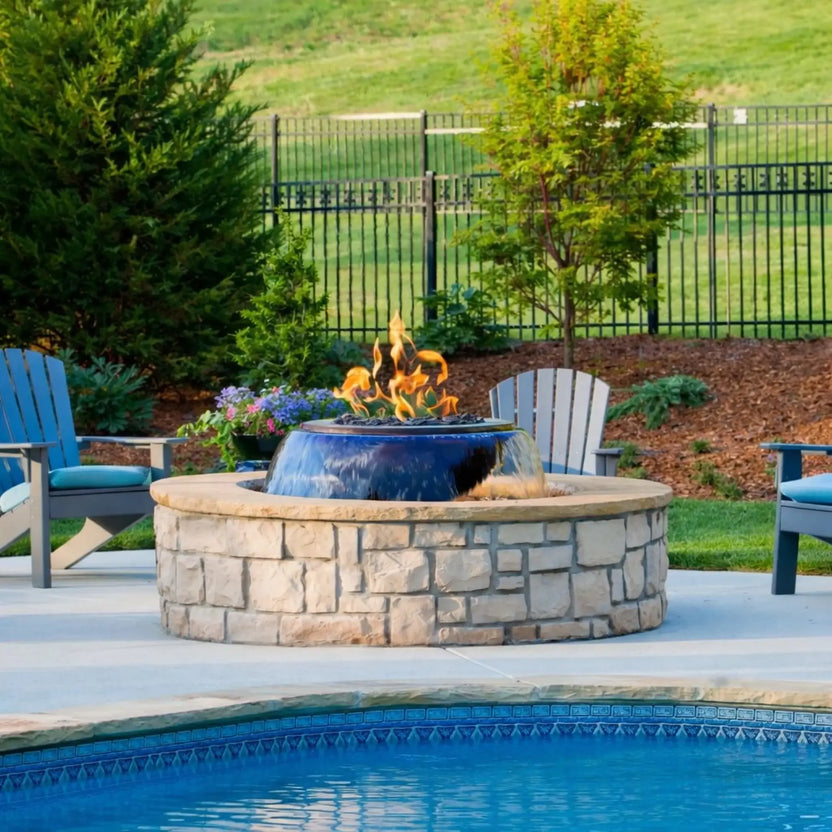 Poolside installation of the Evolution 360 True Fire Water feature within a circular stone enclosure. Flames flicker above the bowl while water flows around it, with Adirondack chairs, landscaped greenery, and a fenced yard creating a polished outdoor living space.