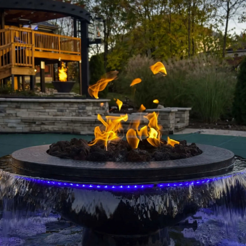 Close-up lifestyle view of the Evolution 360 True Fire Water feature with vibrant flames rising over lava rock while water cascades around the bowl. Blue LED lighting illuminates the falling water, and a deck and landscaped backyard appear softly in the background, emphasizing warmth and ambiance.