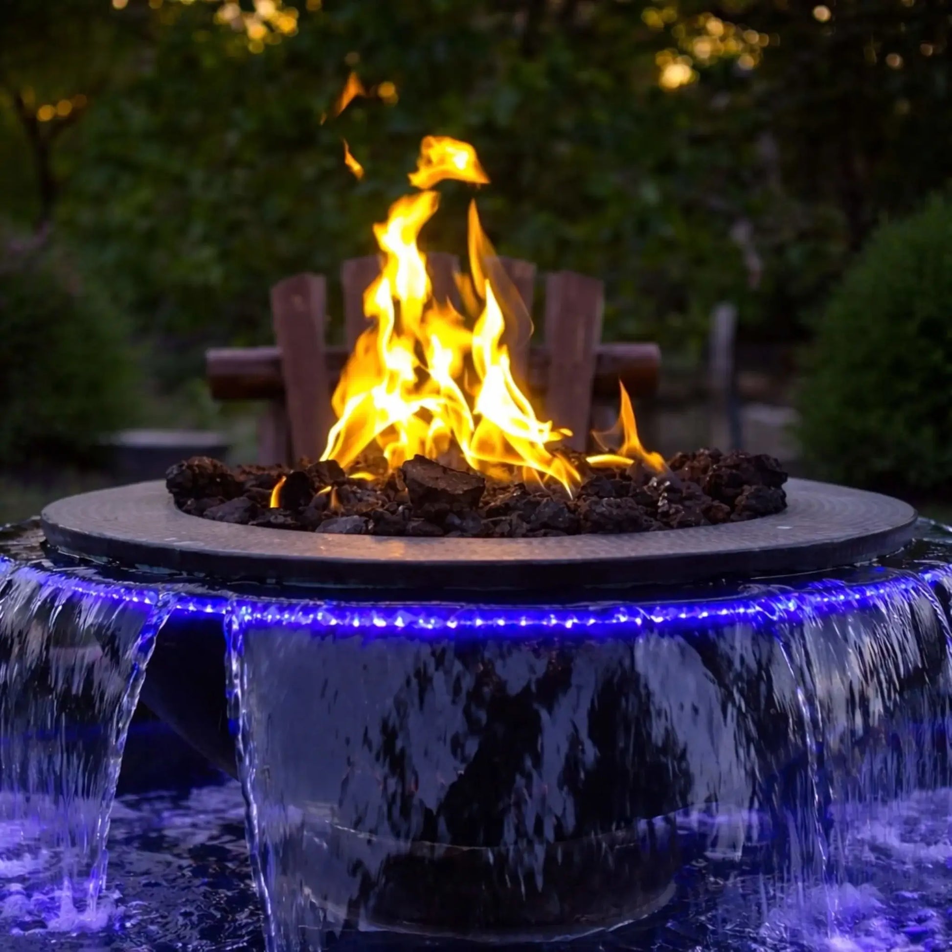Evening close-up of the HPC Evolution 360 fire and water feature with tall, natural flames centered above dark fire media. Water pours from multiple scuppers while blue LED lighting enhances the cascading effect, set against a lush green garden backdrop for a cozy outdoor atmosphere.