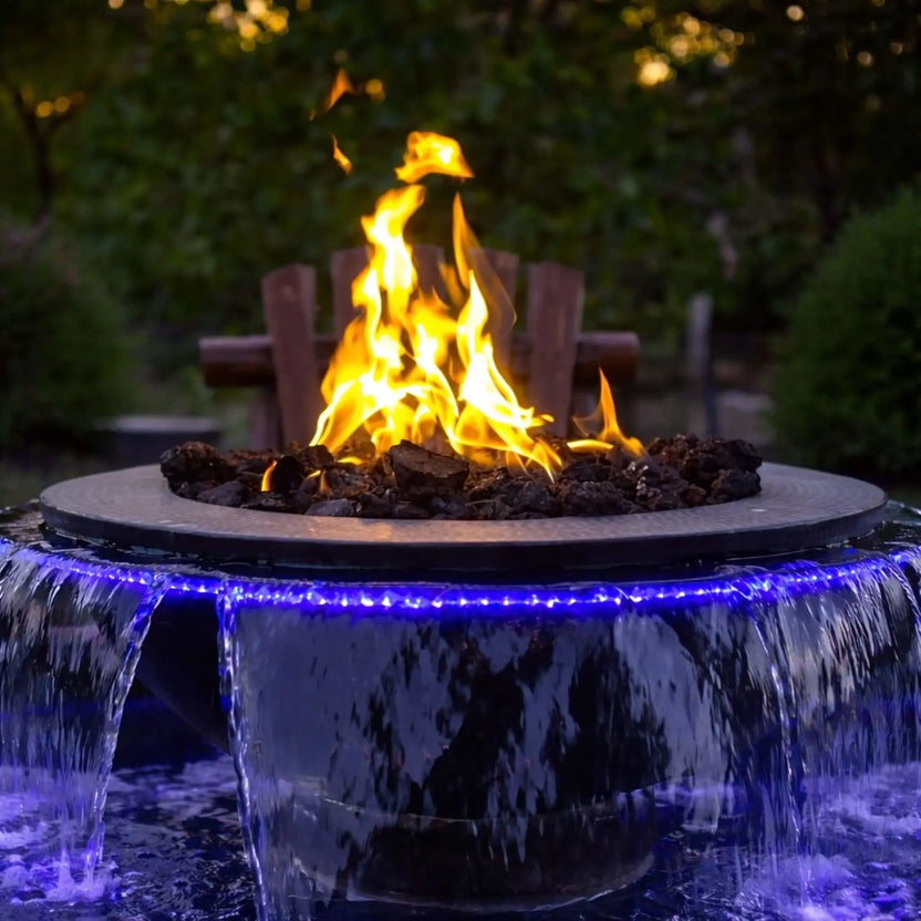 Evening close-up of the HPC Evolution 360 fire and water feature with tall, natural flames centered above dark fire media. Water pours from multiple scuppers while blue LED lighting enhances the cascading effect, set against a lush green garden backdrop for a cozy outdoor atmosphere.