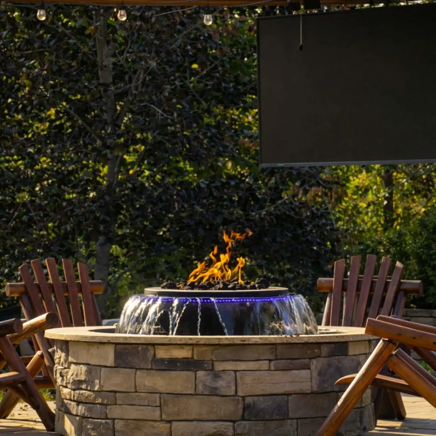Wide patio scene featuring the Evolution 360 True Fire Water feature surrounded by wooden Adirondack chairs under a pergola with string lights. A mounted outdoor screen hangs above, showcasing a complete entertainment space centered around the fire and water feature.