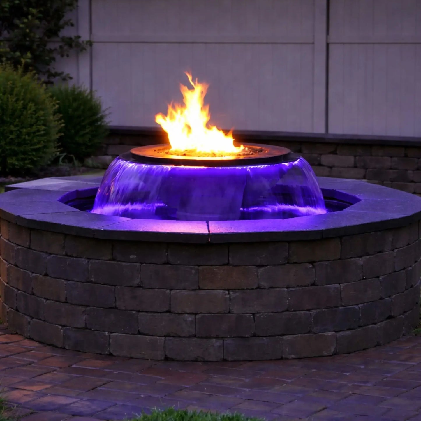 Nighttime view of the Evolution 360 fire and water feature glowing with purple-blue LED lighting beneath a strong flame. The circular brick enclosure contrasts with the illuminated waterfall, creating a dramatic backyard focal point against a fence and landscaped greenery.