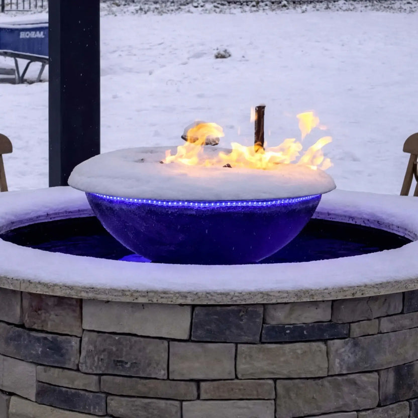 Winter scene featuring the Evolution 360 True Fire Water feature covered in fresh snow, with flames still burning above the bowl. Blue LED lighting glows beneath the icy edge while the surrounding stone enclosure and snowy yard emphasize year-round usability.