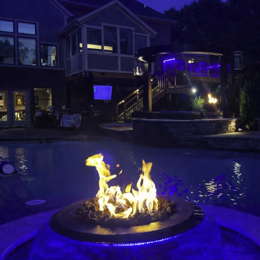 Backyard gathering scene with two women seated in wooden chairs around the Evolution 360 fire and water feature. Flames flicker above the bowl while water flows below, framed by landscaped greenery, stone patio pavers, and a relaxed outdoor seating arrangement.