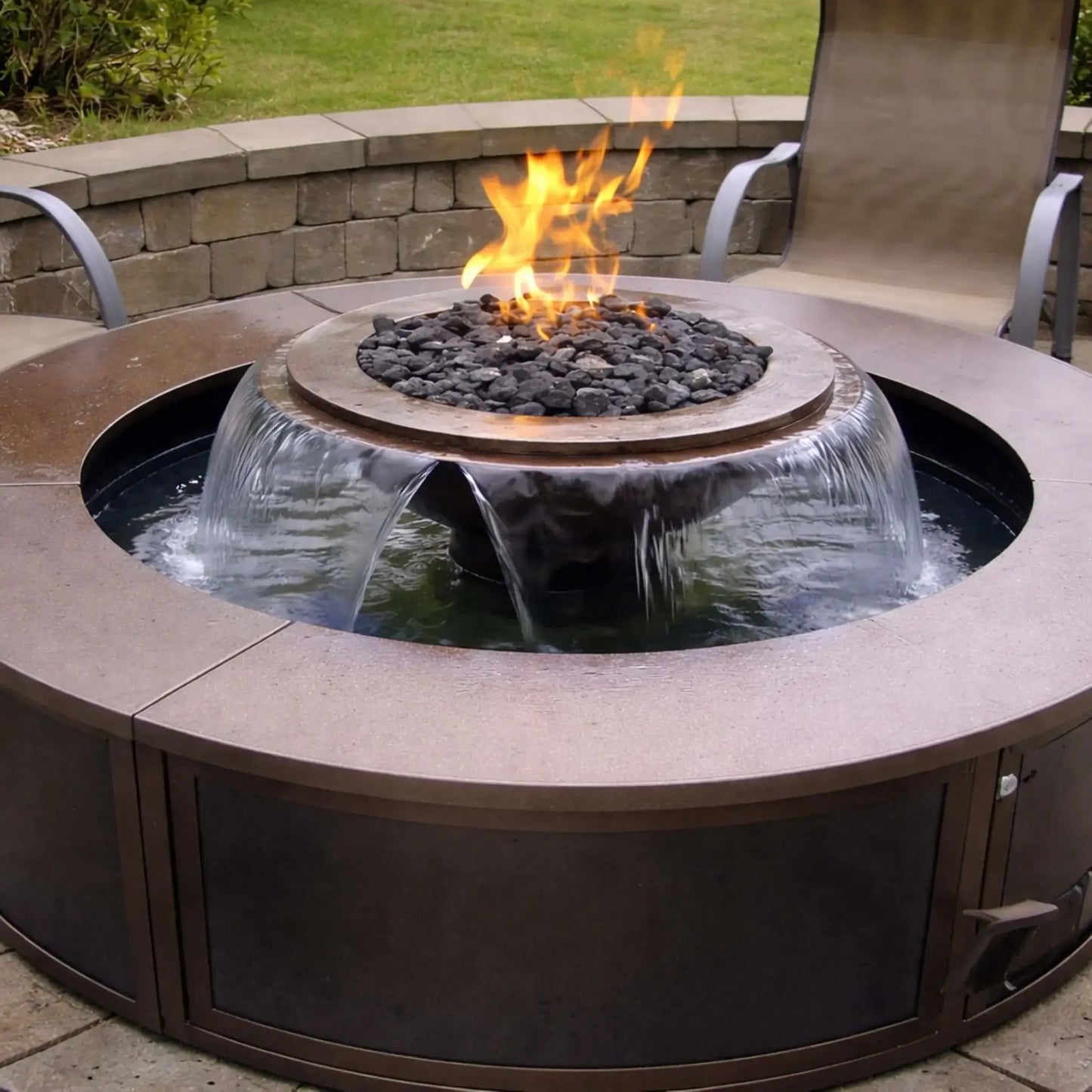 Modern outdoor fire table setup featuring the Evolution 360 True Fire Water system with a circular metal enclosure. Flames burn over black fire media while water spills evenly from multiple scuppers into the surrounding basin, with patio seating and stone wall in the background.
