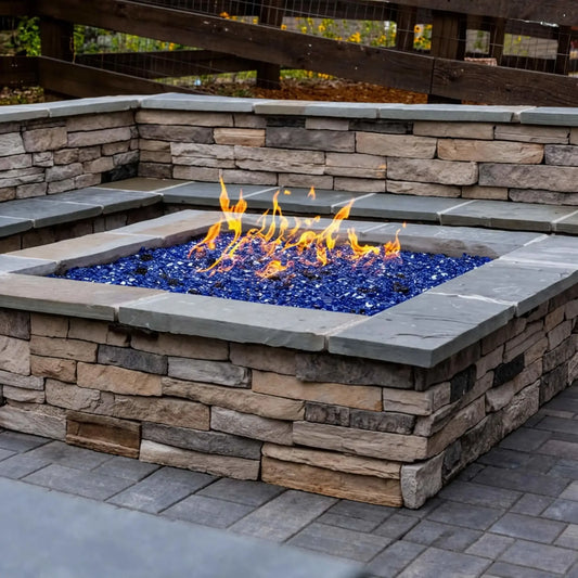 Built-in square fire pit made from stacked stone with a smooth cap, filled with vibrant blue fire glass and producing even, dancing flames. Integrated into a paved patio with matching stone seating walls, this setup highlights a stylish and durable outdoor gathering space with strong visual contrast and texture.