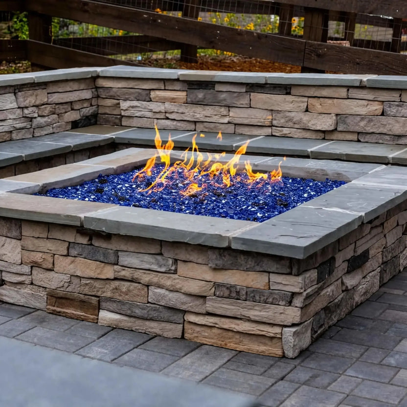 Built-in square fire pit made from stacked stone with a smooth cap, filled with vibrant blue fire glass and producing even, dancing flames. Integrated into a paved patio with matching stone seating walls, this setup highlights a stylish and durable outdoor gathering space with strong visual contrast and texture.