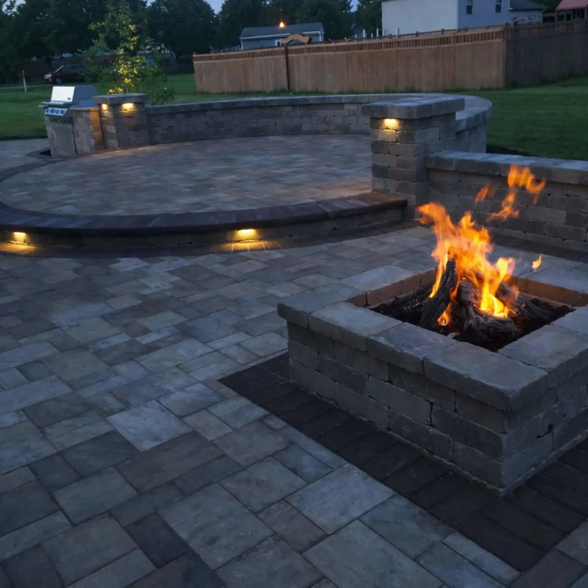 Spacious backyard patio at dusk featuring a square stone fire pit with natural-looking logs and bright flames. Surrounded by large paver stones, curved retaining walls, and built-in landscape lighting, the outdoor living area includes a grill station and open space for entertaining in a suburban setting.