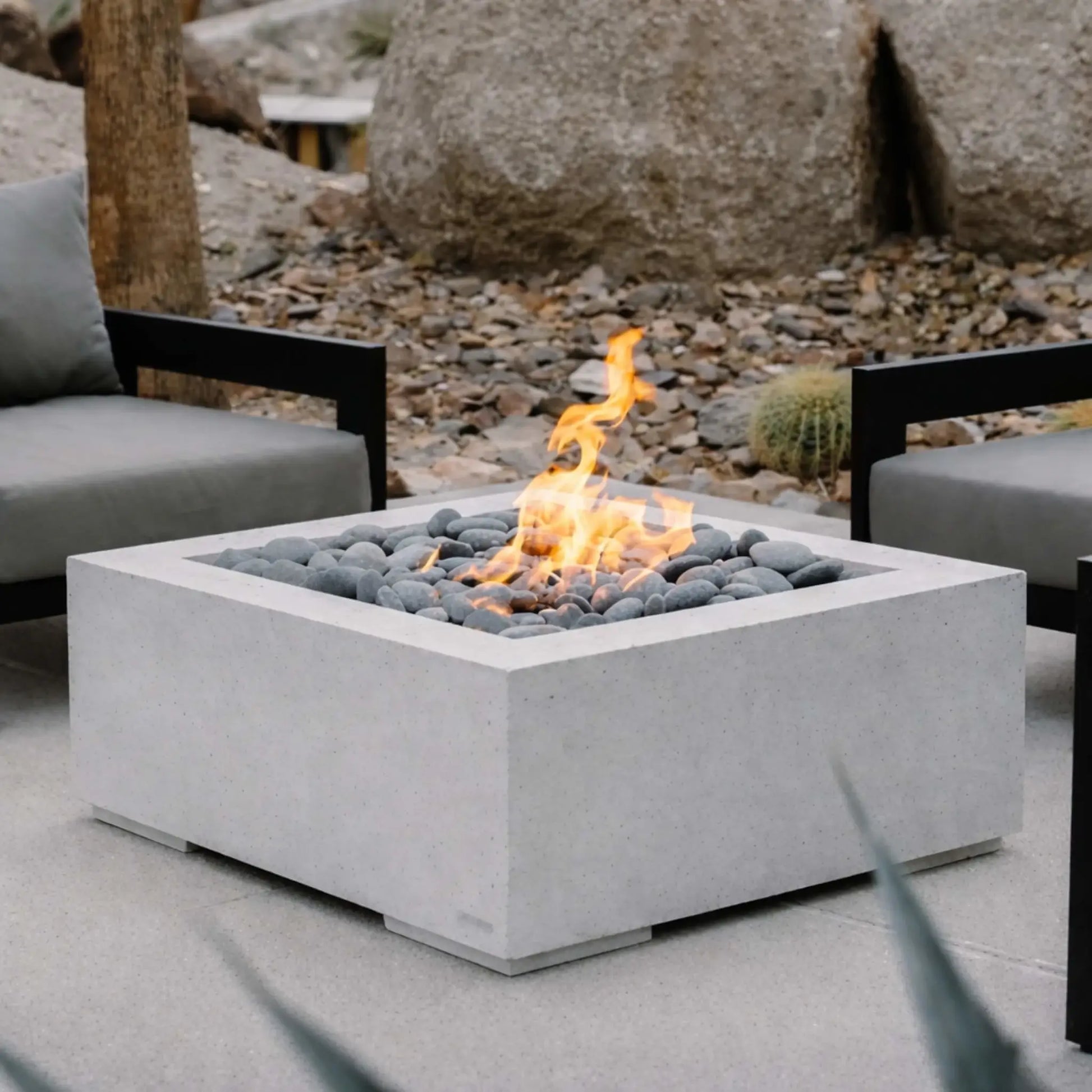 Modern square fire pit with a smooth light concrete finish, filled with rounded gray fire stones and a clean-burning flame. Positioned between two minimalist outdoor chairs, the setup is surrounded by natural rock landscaping and desert-style plants, creating a calm and contemporary outdoor lounge space.