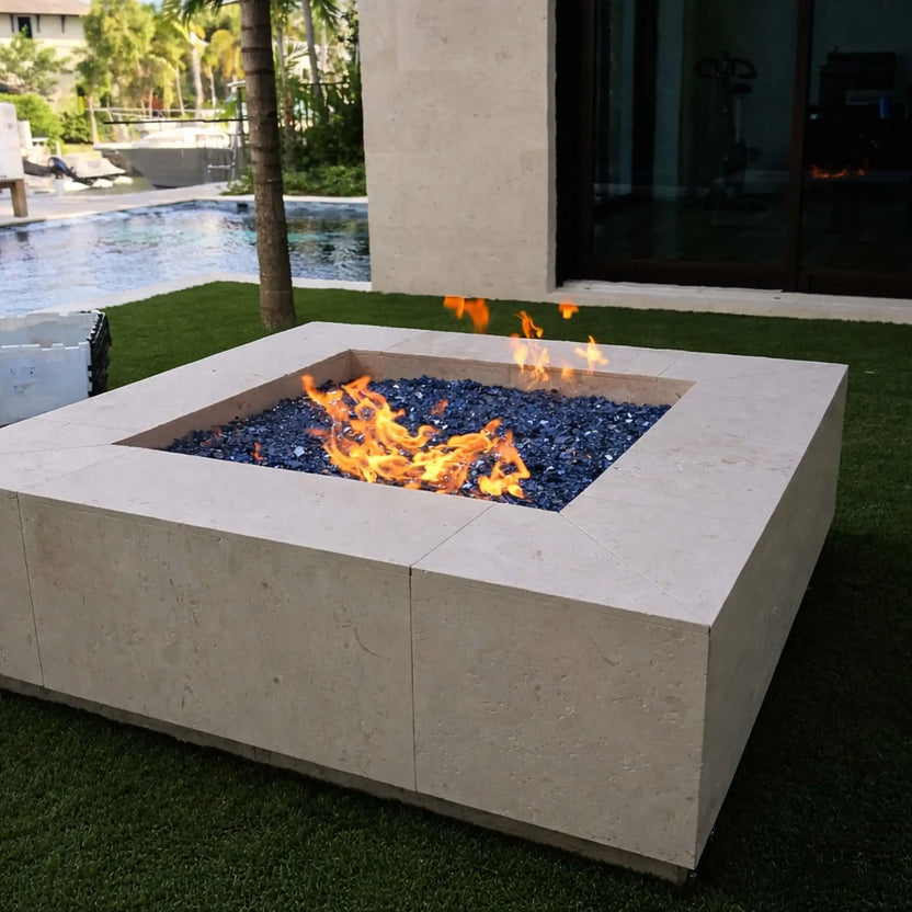 Large square fire pit constructed from light beige stone, filled with dark reflective fire glass and active flames. Located in a luxury backyard near a swimming pool and waterfront, the design blends modern architecture with tropical landscaping, offering a high-end outdoor entertaining environment.