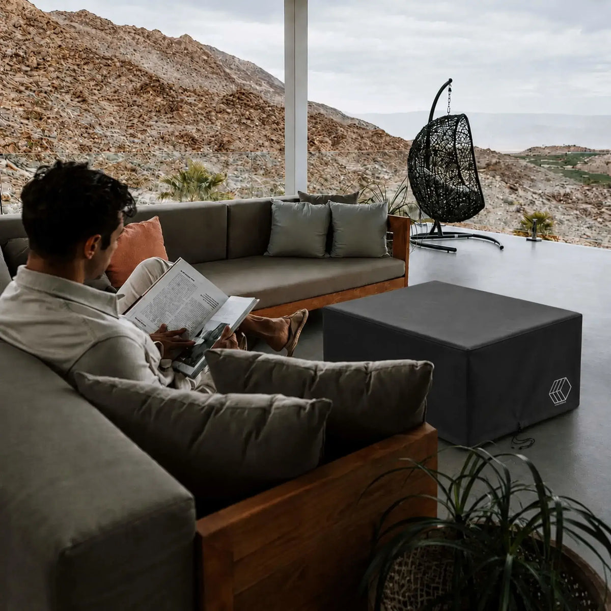 HPC Fire Hearth Products Controls GFRC Concrete Alea 32 x 32 Weather Cover displayed in a luxury outdoor living space. The black weather-resistant cover protects a square Alea fire pit table while blending seamlessly with modern patio furniture, including cushioned sectional seating and a hanging chair. Set against a scenic desert mountain backdrop, this lifestyle image highlights how the Alea 32 x 32 Weather Cover safeguards outdoor fire features while maintaining a refined contemporary aesthetic.