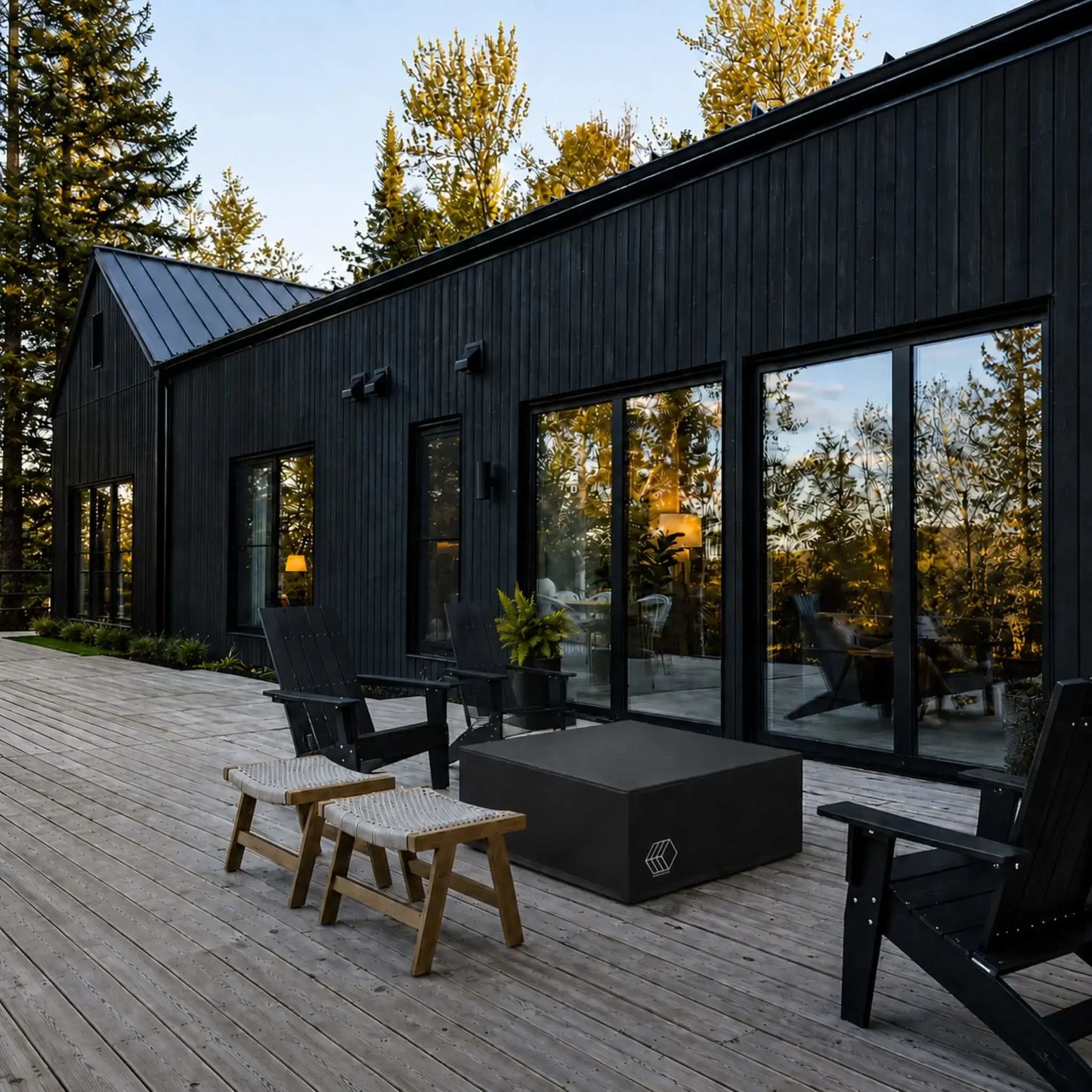 HPC Fire Hearth Products Controls GFRC Concrete Alea 40 x 40 Weather Cover displayed on a spacious contemporary wood deck outside a modern black-clad home with expansive glass windows. The fitted black protective cover conceals and protects a square Alea fire pit table between Adirondack chairs and outdoor seating. This lifestyle setting demonstrates the cover’s ability to preserve outdoor fire features from rain, snow, debris, UV damage, and changing weather conditions while complementing modern Patio.