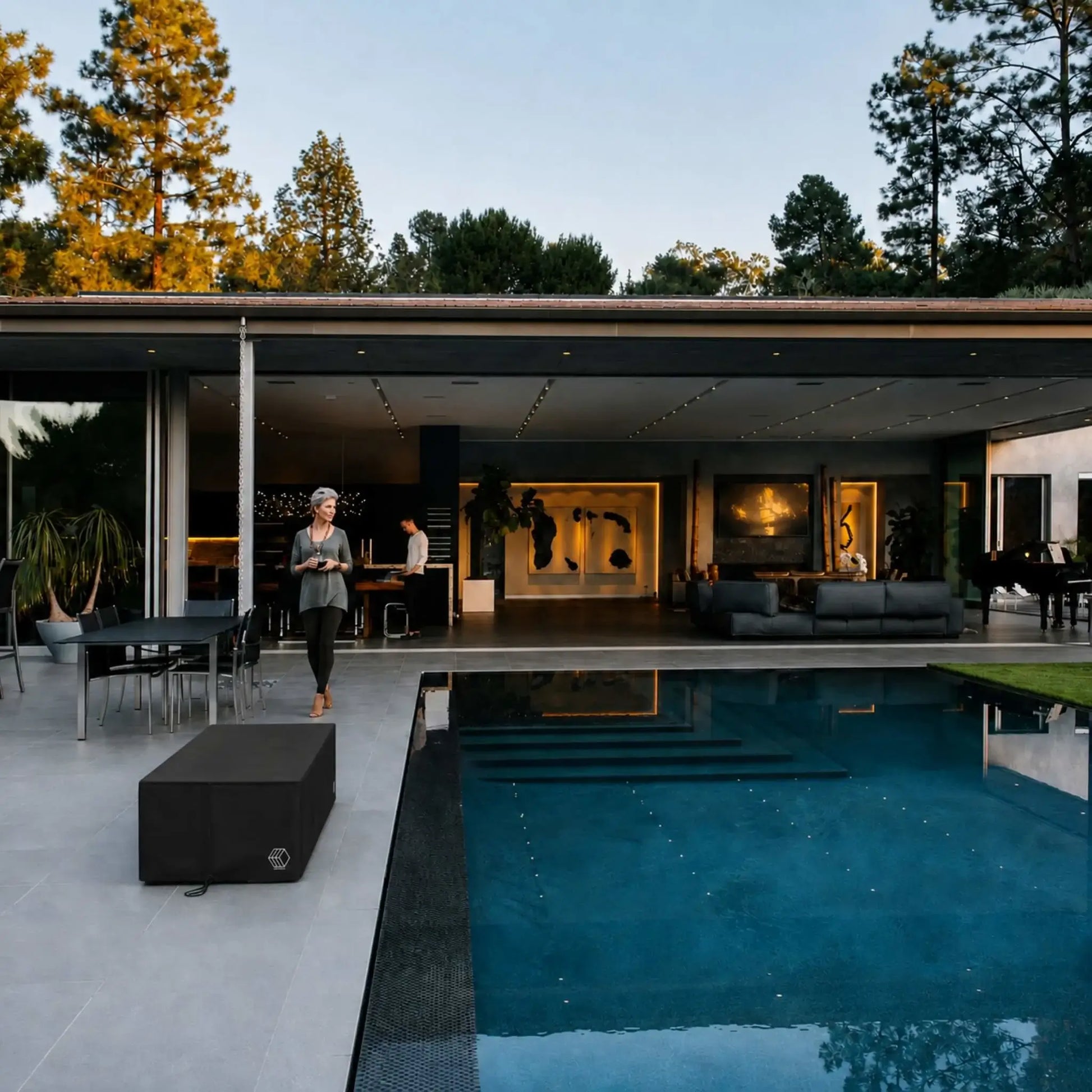 HPC Fire & Hearth Products Controls GFRC Concrete Avera 48 x 26 Weather Cover displayed beside a modern luxury swimming pool at a contemporary outdoor living space. The black rectangular weather cover protects a fire pit table while blending seamlessly with upscale patio furnishings, expansive open-air architecture, and poolside entertainment areas, demonstrating how the cover preserves outdoor fire features from the elements without compromising design aesthetics.
