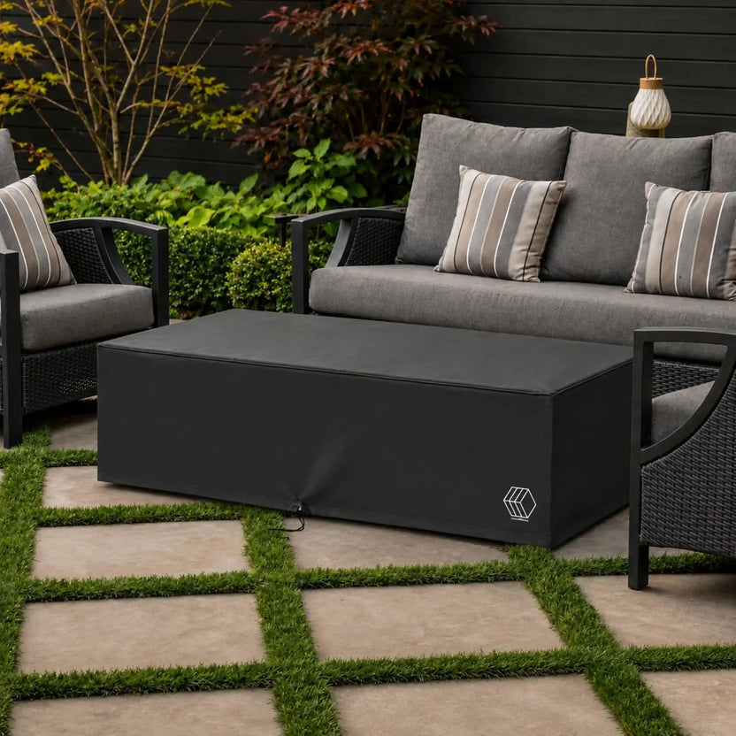 HPC Fire Hearth Products Controls GFRC Concrete Avera 60 x 26 Weather Cover Lifestyle 1 image showing the fitted black weather cover installed over an Avera fire pit in an outdoor seating area. Surrounded by gray cushioned patio furniture, decorative landscaping, and modern paver walkways, the cover provides reliable protection from weather and debris while maintaining a clean, sophisticated appearance within the outdoor living space.