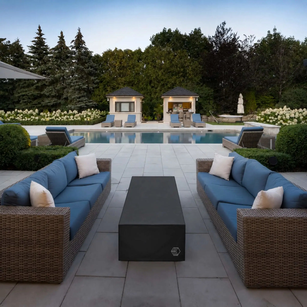 HPC Fire Hearth Products Controls GFRC Concrete Avera 60 x 26 Weather Cover Lifestyle 2 image featuring the covered fire pit positioned between two blue-cushioned wicker sofas beside a luxury swimming pool. The sleek black weather cover preserves the fire pit’s condition while blending seamlessly into an upscale outdoor entertainment setting complete with poolside seating, manicured landscaping, cabanas, and expansive patio surfaces.