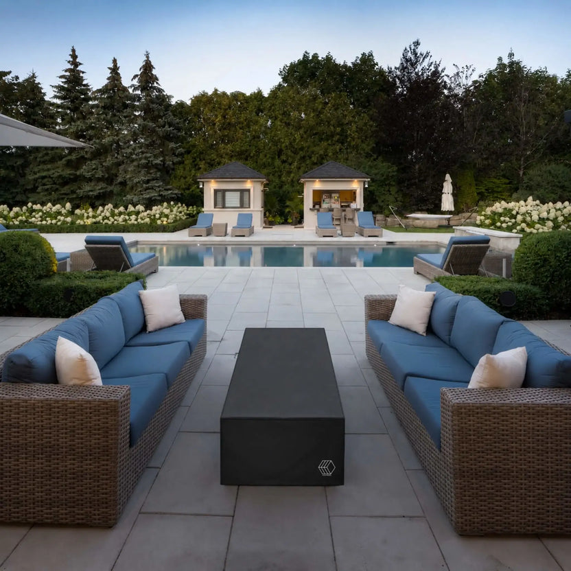 HPC Fire Hearth Products Controls GFRC Concrete Avera 60 x 26 Weather Cover Lifestyle 2 image featuring the covered fire pit positioned between two blue-cushioned wicker sofas beside a luxury swimming pool. The sleek black weather cover preserves the fire pit’s condition while blending seamlessly into an upscale outdoor entertainment setting complete with poolside seating, manicured landscaping, cabanas, and expansive patio surfaces.