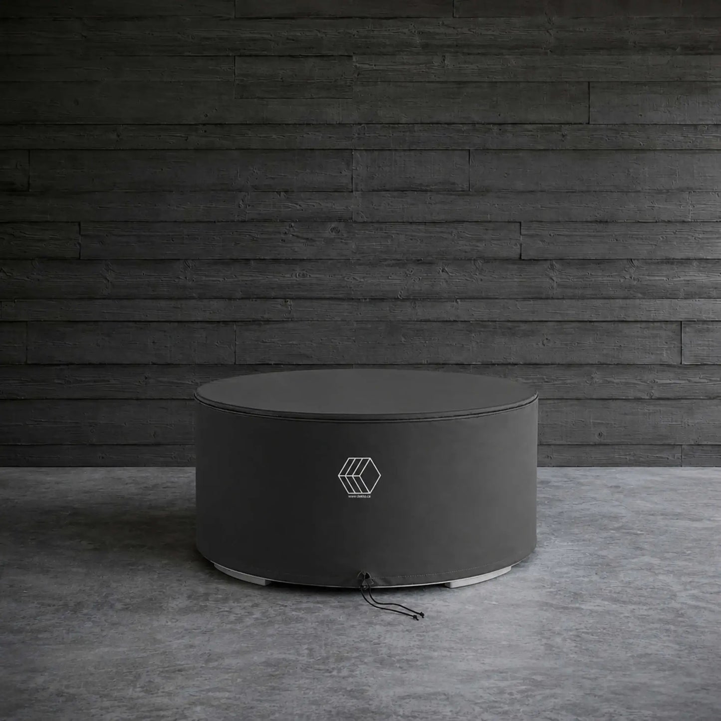 HPC Fire Hearth Products GFRC Concrete Belmont Weather Cover displayed in a lifestyle setting against a dark wood plank wall and concrete floor. The round black protective cover is fitted over a circular Belmont fire pit, showcasing its sleek, low-profile design and weather-resistant construction. Reinforced stitching and a secure drawstring closure help shield the fire feature from rain, dust, leaves, and other outdoor elements.