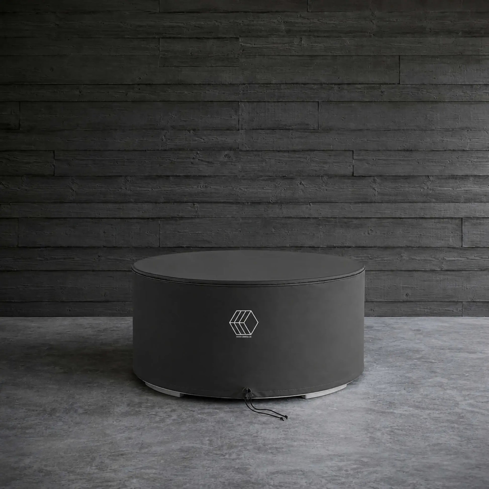 HPC Fire Hearth Products GFRC Concrete Belmont Weather Cover displayed in a lifestyle setting against a dark wood plank wall and concrete floor. The round black protective cover is fitted over a circular Belmont fire pit, showcasing its sleek, low-profile design and weather-resistant construction. Reinforced stitching and a secure drawstring closure help shield the fire feature from rain, dust, leaves, and other outdoor elements.
