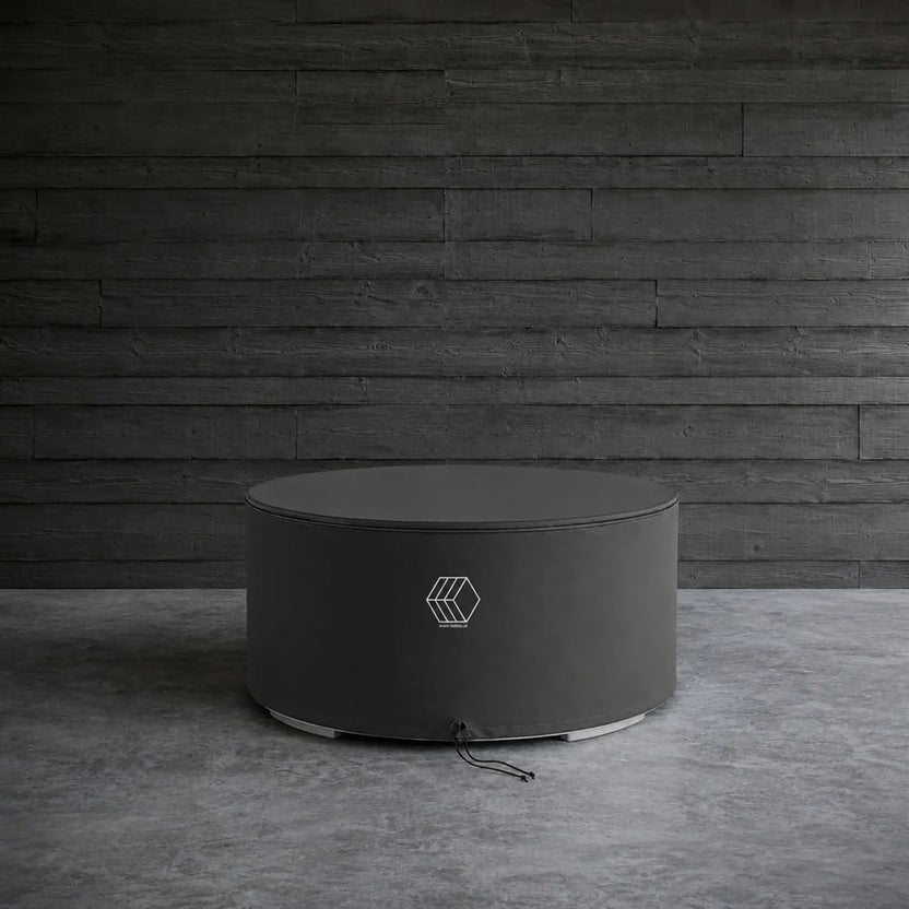 HPC Fire Hearth Products GFRC Concrete Belmont Weather Cover displayed in a lifestyle setting against a dark wood plank wall and concrete floor. The round black protective cover is fitted over a circular Belmont fire pit, showcasing its sleek, low-profile design and weather-resistant construction. Reinforced stitching and a secure drawstring closure help shield the fire feature from rain, dust, leaves, and other outdoor elements.